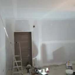 Pokój w trakcie prac wykończeniowych: ściany przygotowane pod malowanie, częściowo pokryte gładzią, widoczna drabina, narzędzia i materiały budowlane na podłodze, surowe oświetlenie żarówką.