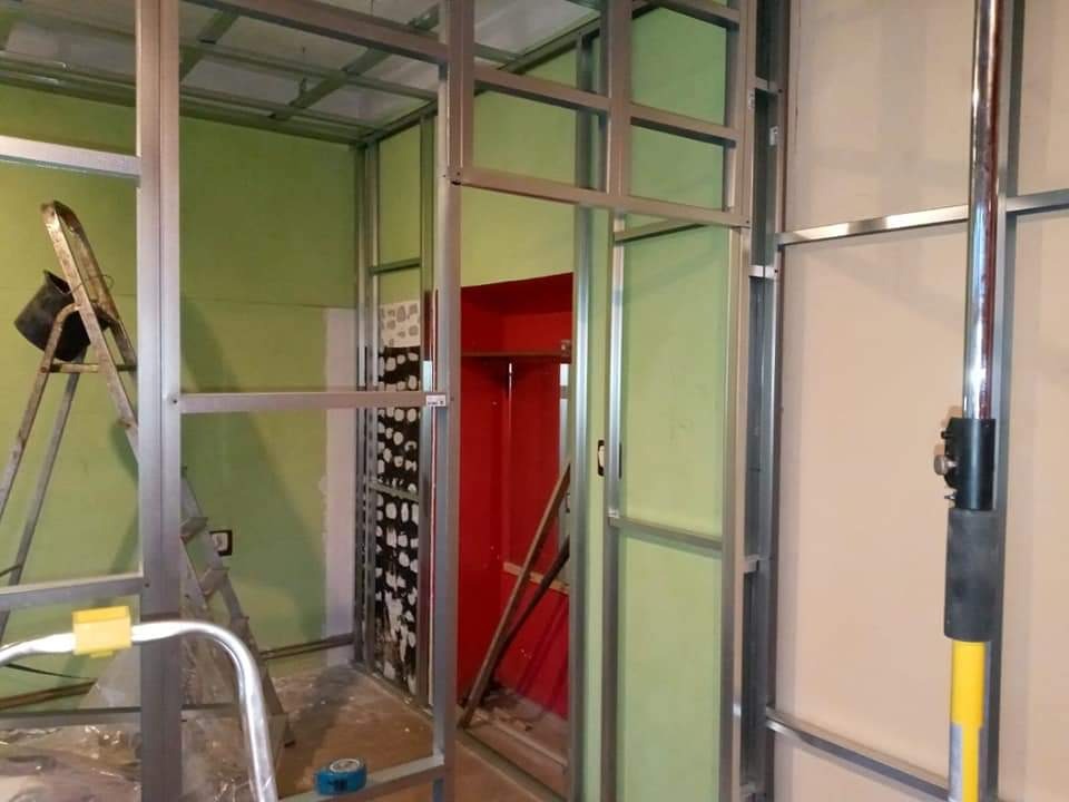 Montaż metalowej konstrukcji pod płyty karton gips: widoczne profile, drabina z wiadrem, częściowo wykończona ściana z instalacją elektryczną, czerwony fragment ściany w głębi.