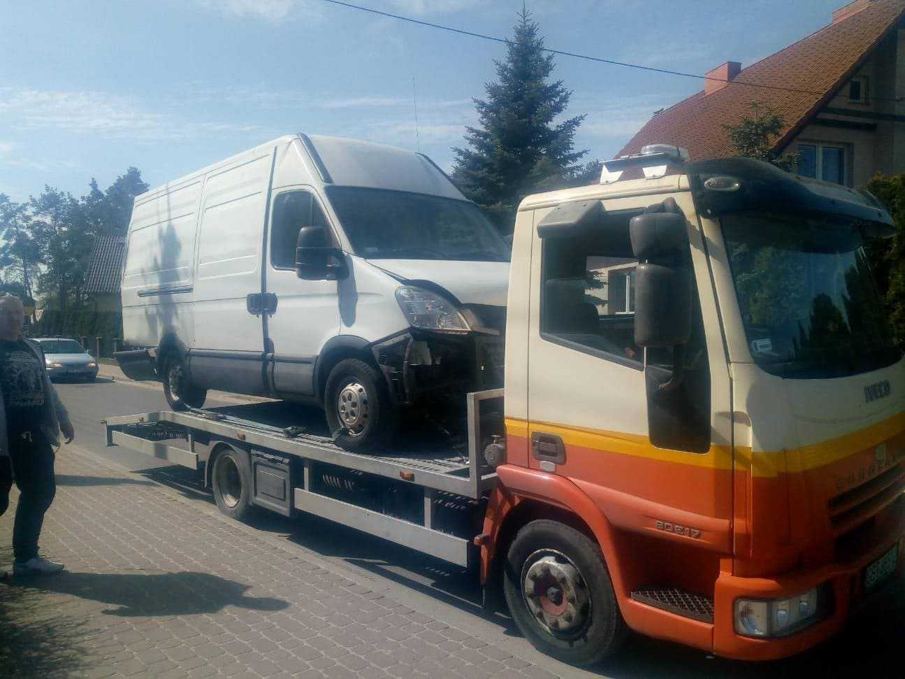 Uszkodzony biały van marki Iveco na platformie pomarańczowo-białej lawety Iveco Cargo 80E17 na tle chodnika i domów.