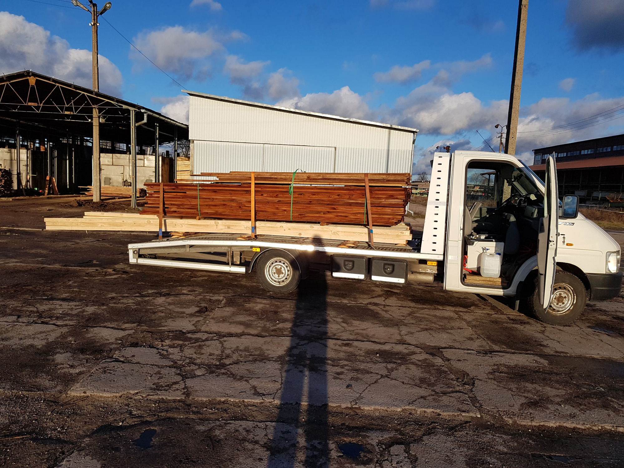 Biała laweta z otwartymi drzwiami kabiny, załadowana drewnem budowlanym, na tle błękitnego nieba z chmurami i zabudowań przemysłowych