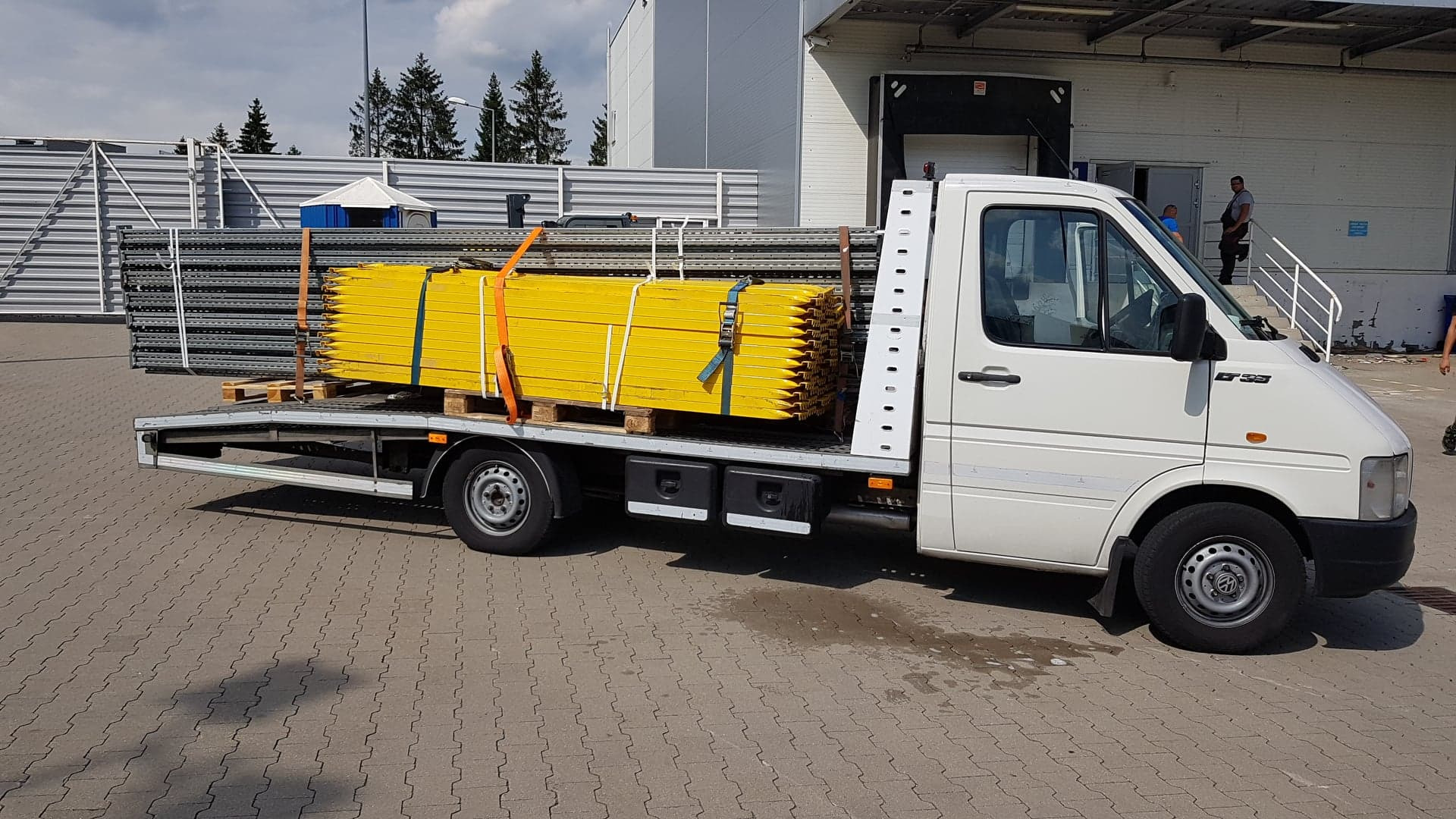 Biała laweta z platformą przewozi paletę z żółtymi i szarymi elementami konstrukcyjnymi, zabezpieczonymi pomarańczowymi i niebieskimi pasami, na tle magazynu z rampą.