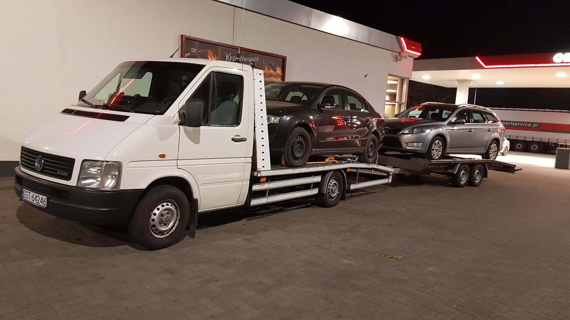 Biała laweta z dwoma samochodami osobowymi na platformie, stojąca na oświetlonym parkingu stacji benzynowej nocą.