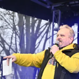 Mężczyzna z mikrofonem na scenie, ubrany w żółtą bluzę z kapturem i czarną kamizelkę, wskazuje palcem trzymając białą kartkę, w tle oświetlenie sceniczne i drzewa.