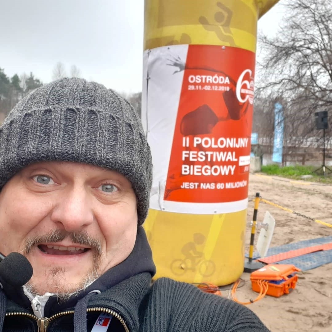 Mężczyzna z mikrofonem na tle bramy startowej II Polonijnego Festiwalu Biegowego w Ostródzie, z widocznym sprzętem nagłośnieniowym.