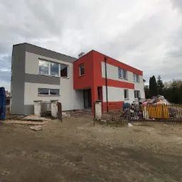 Dwukondygnacyjny budynek z częściowo pomalowaną elewacją w kolorach szarym, białym i czerwonym, widoczny z zewnątrz, z gruzem i narzędziami budowlanymi na podwórku, pod pochmurnym niebem.