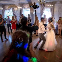 Filmowanie pierwszego tańca pary młodej podczas wesela, z widocznym kamerzystą używającym stabilizatora obrazu, goście w tle klaszczą.