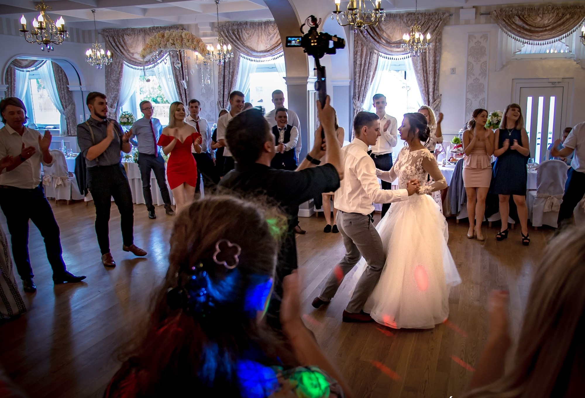 Filmowanie pierwszego tańca pary młodej podczas wesela, z widocznym kamerzystą używającym stabilizatora obrazu, goście w tle klaszczą.