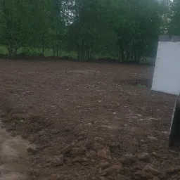 Przygotowany teren pod budowę, świeżo przekopana ziemia z widocznymi bryłami, w tle rząd drzew i fragment białej płyty.