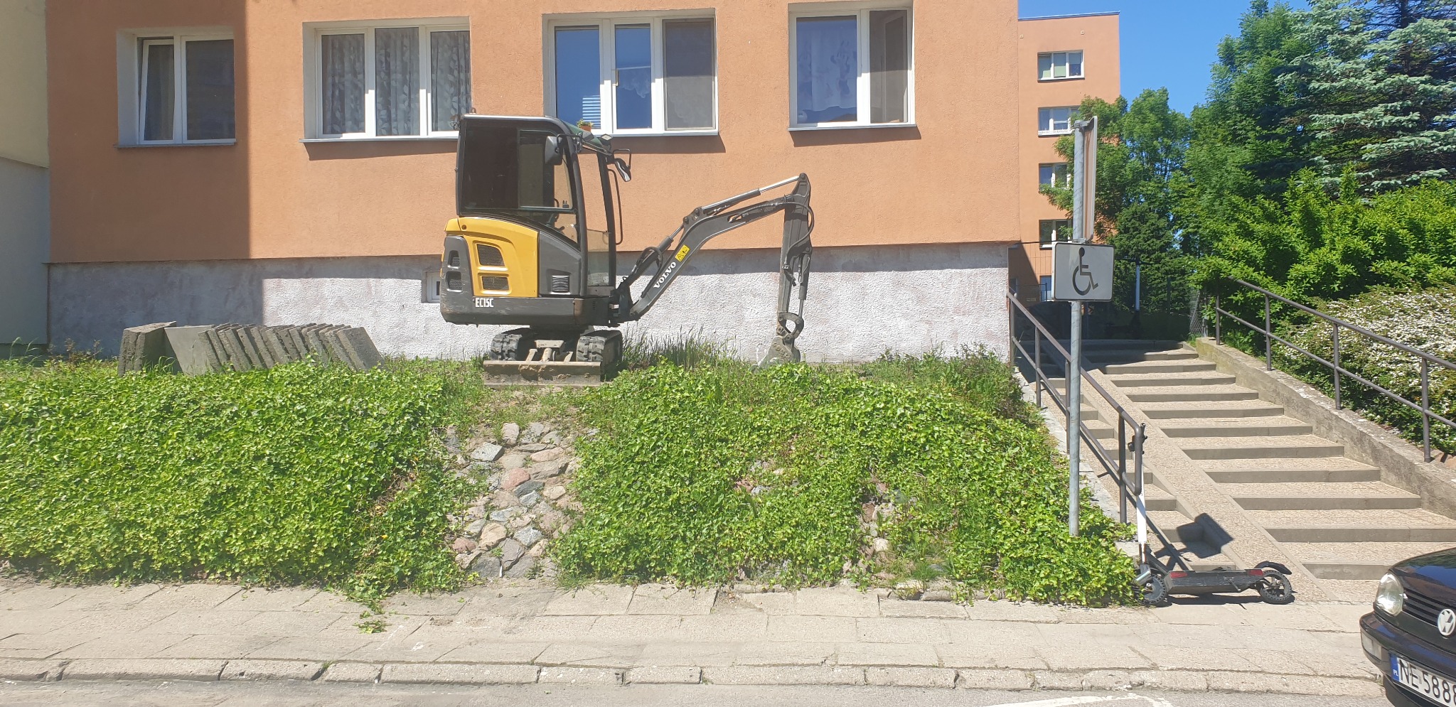 Żółto-czarna mini koparka Volvo stoi na niewielkim wzniesieniu porośniętym zielonym bluszczem, w tle budynek mieszkalny z oknami i schody z podjazdem dla wózków inwalidzkich.