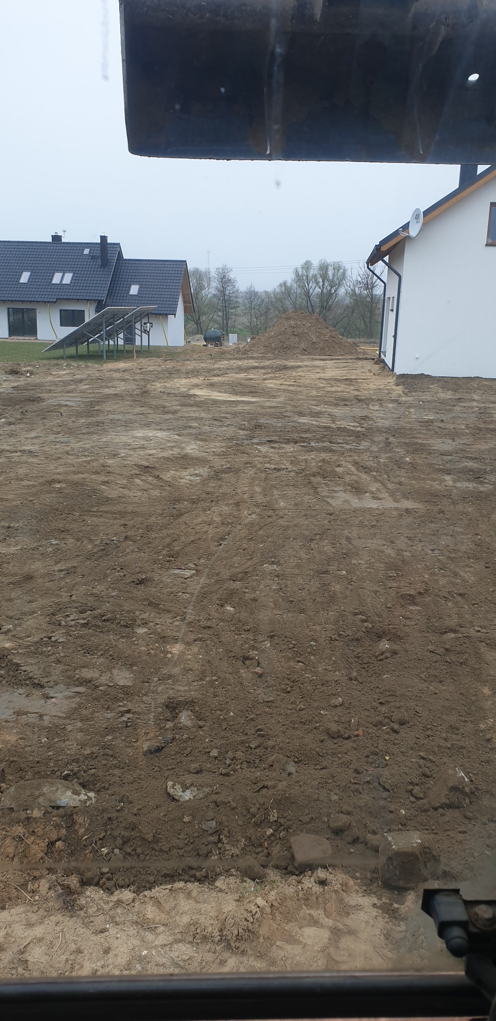 Przygotowany teren pod budowę domu, widoczny z kabiny koparki, z hałdą ziemi w tle i dwoma budynkami mieszkalnymi, jeden z panelami słonecznymi.