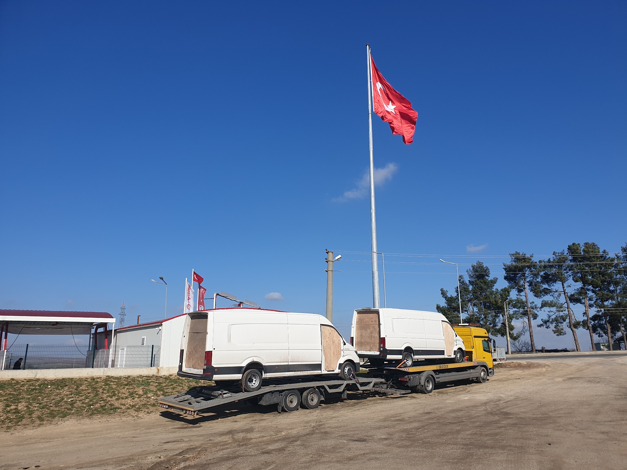 Żółta laweta przewozi dwa białe busy na tle błękitnego nieba i powiewającej tureckiej flagi.