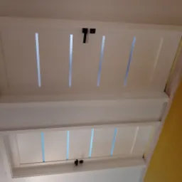 Dwie białe, prostokątne klapy rewizyjne z czarnymi zamkami, umieszczone w suficie, z widocznymi szczelinami wentylacyjnymi i żółtą ścianą w tle.