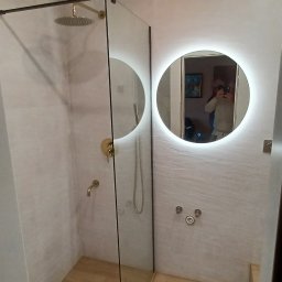 MAT-BUD - Nowoczesna kabina prysznicowa typu walk-in z deszczownicą w kolorze złotym, okrągłe lustro z podświetleniem LED, płytki imitujące drewno na podłodze, szara ściana z subtelną fakturą.