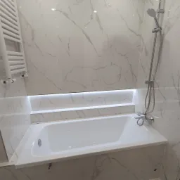 Biała wanna w obudowie z płytek imitujących marmur, podświetlona listwą LED, z chromowaną baterią i słuchawką prysznicową na tle ściany wyłożonej tymi samymi płytkami, obok grzejnik drabinkowy.