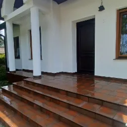 Wejście do domu z brązowymi płytkami na schodach i podestach, białe ściany budynku, czarne drzwi wejściowe, kolumny podtrzymujące zadaszenie.