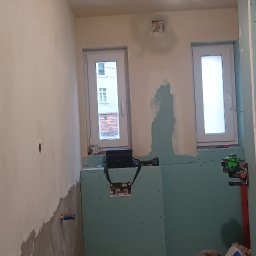 MAT-BUD - Łazienka w trakcie remontu: ściany z płyt karton-gips, częściowo położona gładź, widoczne okna, instalacja sanitarna i pracownik w kurtce podczas pracy.