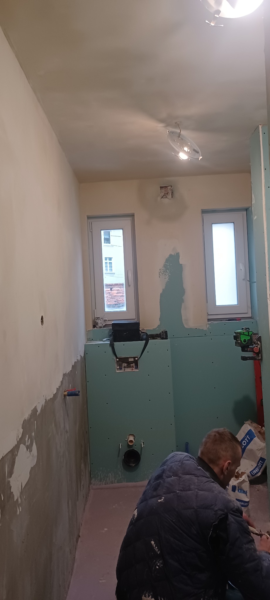 Łazienka w trakcie remontu: ściany z płyt karton-gips, częściowo położona gładź, widoczne okna, instalacja sanitarna i pracownik w kurtce podczas pracy.