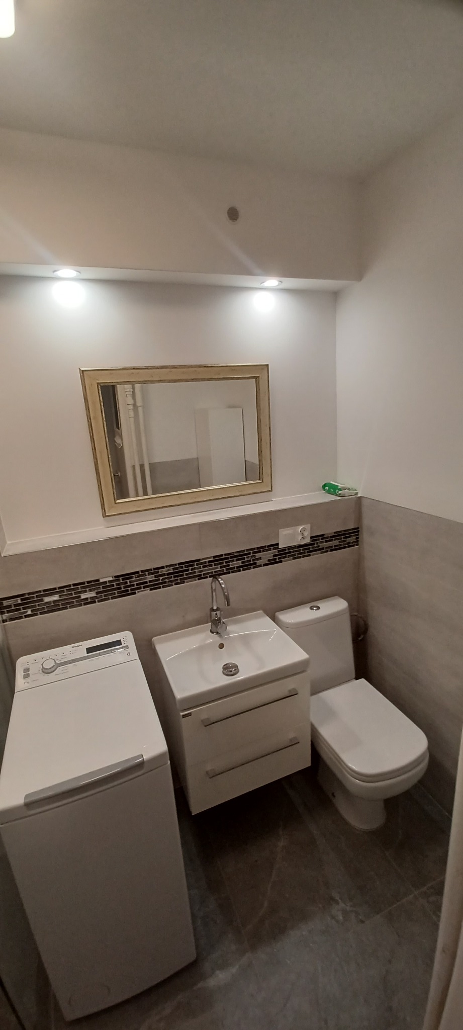 Wnętrze łazienki z pralką, umywalką z szafką i toaletą, wykończone płytkami w odcieniach szarości, z lustrem w ozdobnej ramie i punktowym oświetleniem sufitowym.