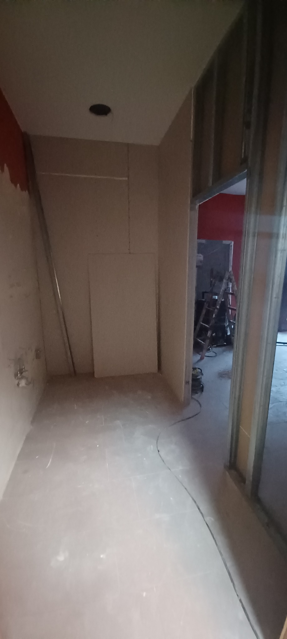 Wnętrze w trakcie prac: ściany z płyt kartonowo-gipsowych, widoczne profile metalowe, instalacja elektryczna w ścianie, surowa podłoga, w tle drabina i czerwona ściana w innym pomieszczeniu.