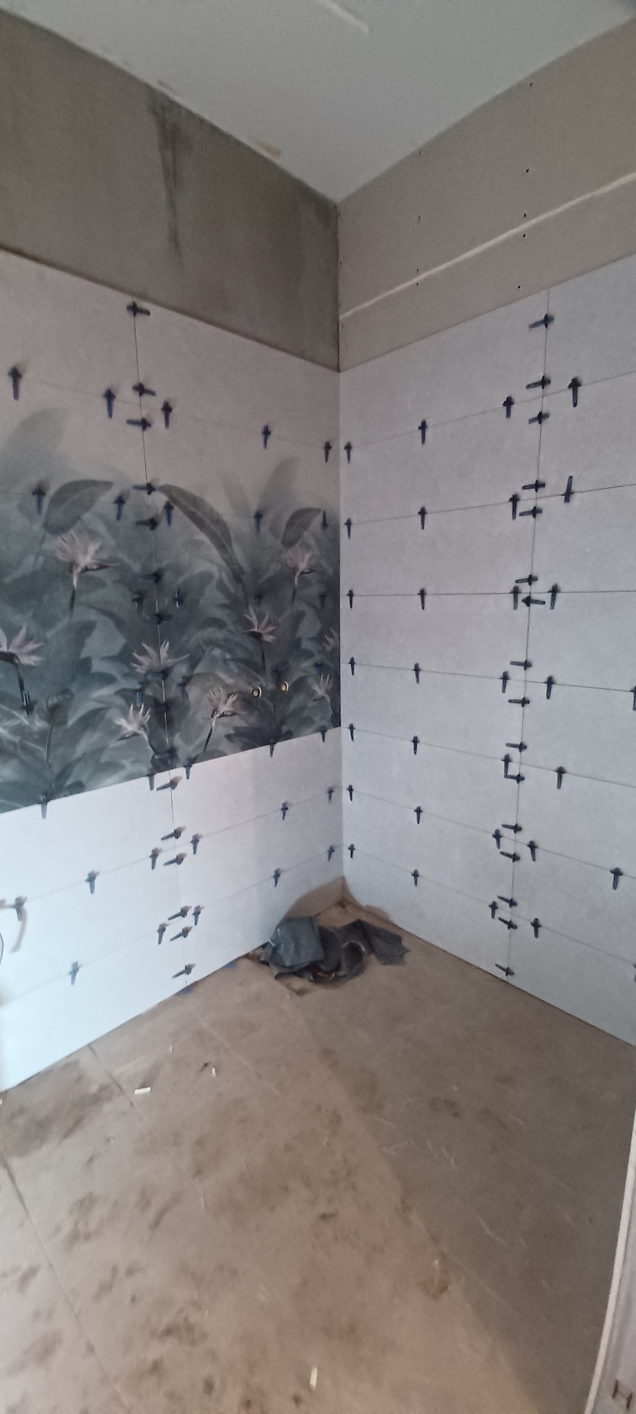 Świeżo ułożone, jasne płytki z motywem roślinnym na ścianie łazienki, z widocznymi krzyżykami dystansowymi i poziomowaniem, podłoga częściowo zakryta folią.
