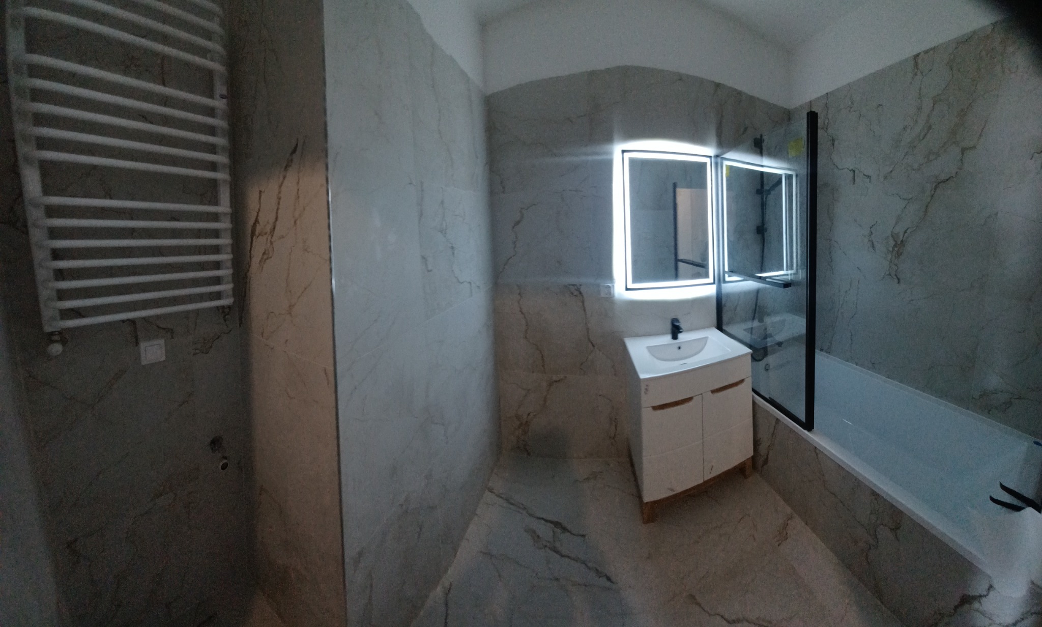 Wykończona łazienka z marmurowymi płytkami, podświetlanym lustrem, grzejnikiem drabinkowym i kabiną prysznicową z czarnymi profilami.