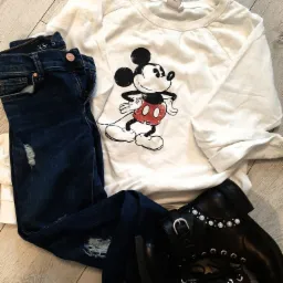 Zestaw odzieży damskiej: biała bluza z nadrukiem Myszki Miki, ciemne jeansy z przetarciami i czarne botki z ozdobnymi perłami i sznurowaniem, ułożone na drewnianej podłodze.