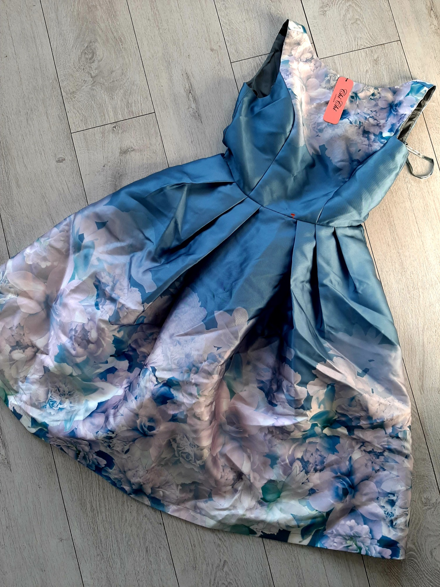 Niebieska, satynowa sukienka z motywem kwiatowym, leżąca na panelach podłogowych. Widoczna metka z nazwą marki.