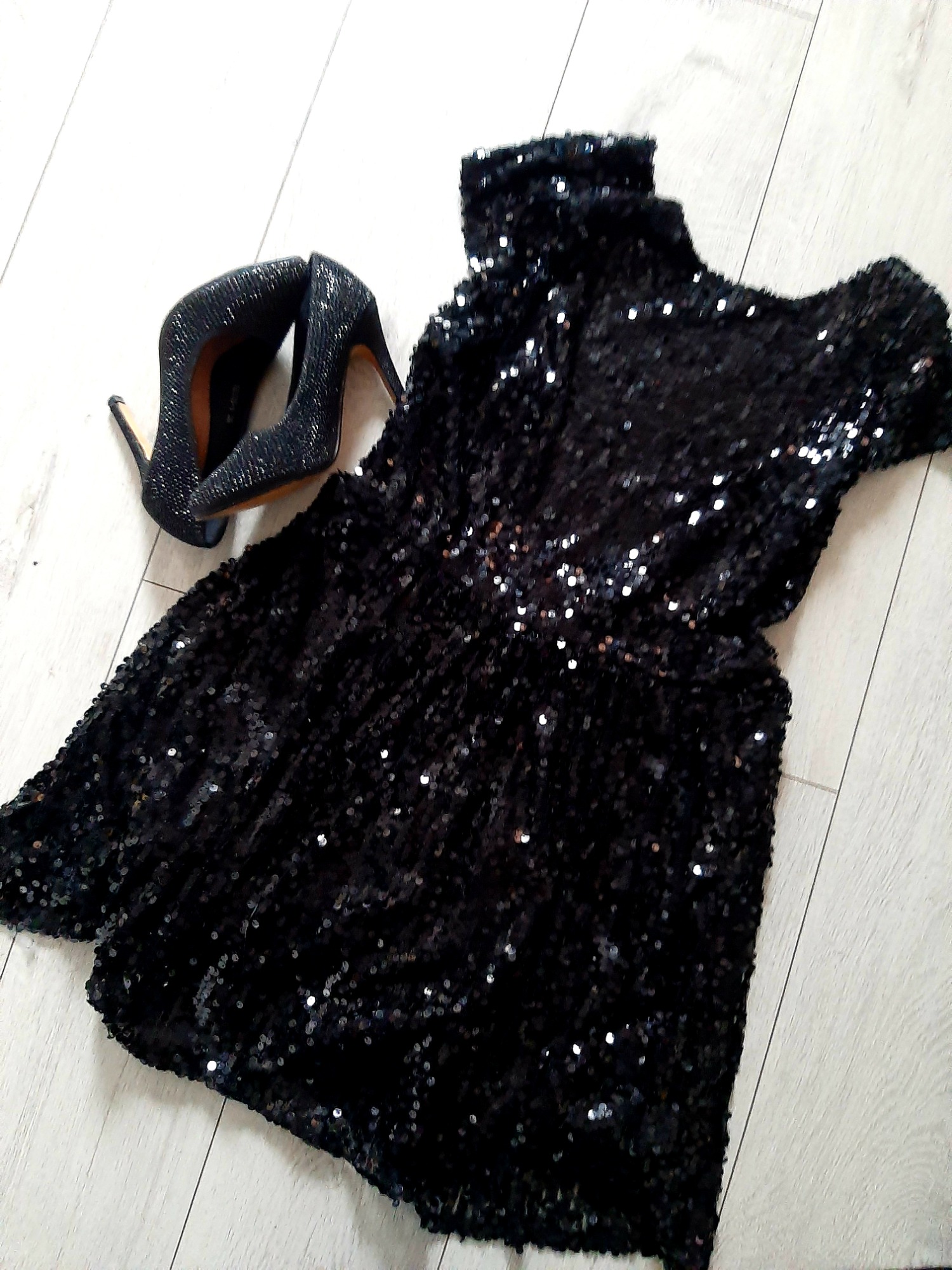 Czarna, cekinowa sukienka z golfem i czarne, brokatowe szpilki na jasnym, drewnianym tle.