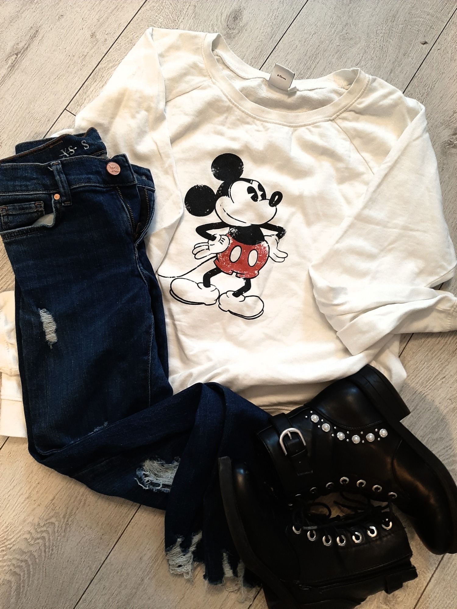 Zestaw odzieży damskiej: biała bluza z nadrukiem Myszki Miki, ciemne jeansy z przetarciami i czarne botki z ozdobnymi perłami i sznurowaniem, ułożone na drewnianej podłodze.
