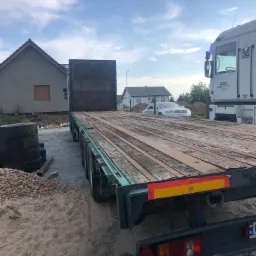 Pusta naczepa niskopodwoziowa z drewnianą podłogą na placu budowy, widoczna sterta opon i budynek w tle.