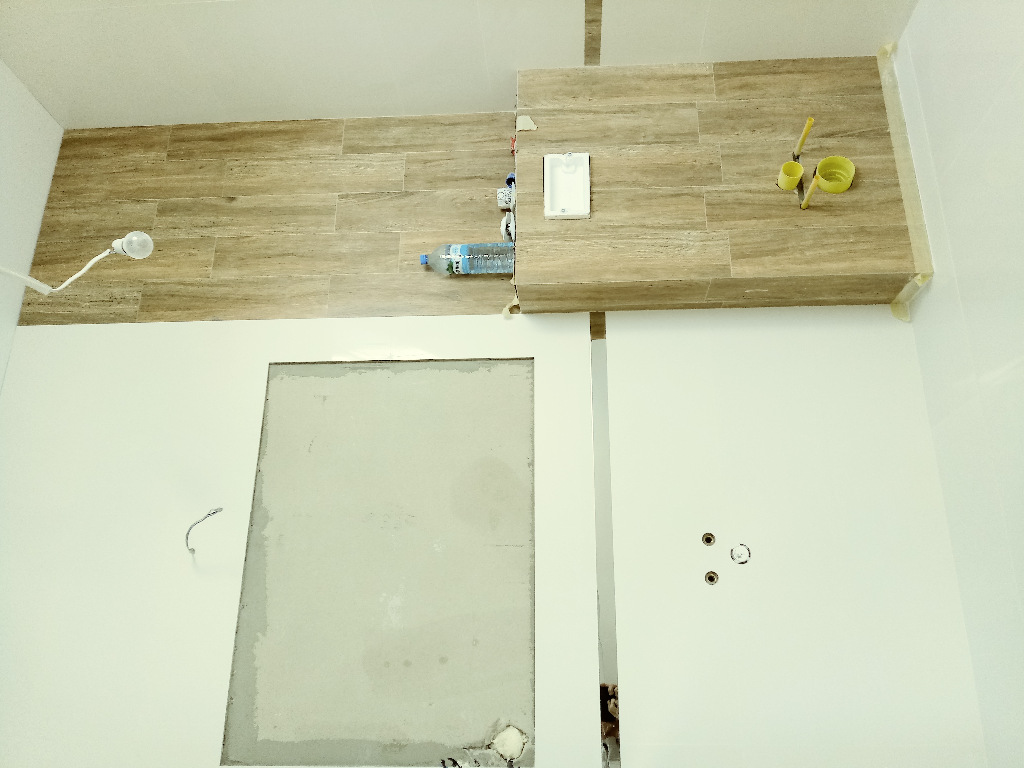 Niedokończona łazienka z częściowo ułożonymi płytkami imitującymi drewno, widoczne narzędzia, butelka wody i otwory w ścianie.