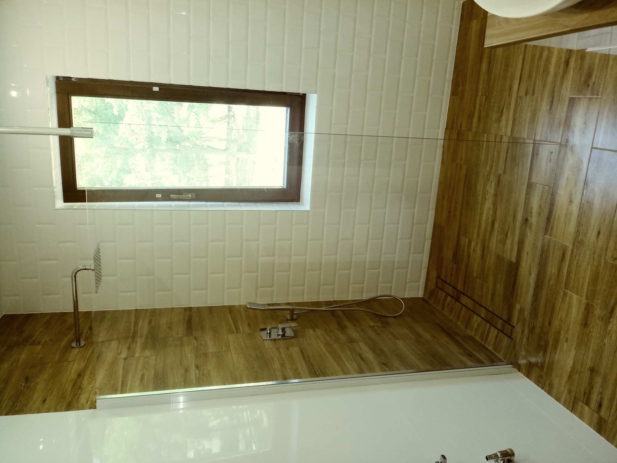 Nowoczesna łazienka z kabiną prysznicową, białe płytki imitujące cegłę na ścianach, drewnopodobna podłoga, okno z widokiem na zieleń.