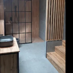 Nowoczesna łazienka na poddaszu z kabiną prysznicową typu loft, umywalką nablatową i drewnianymi elementami dekoracyjnymi, w tym ażurową ścianką działową i schodami.