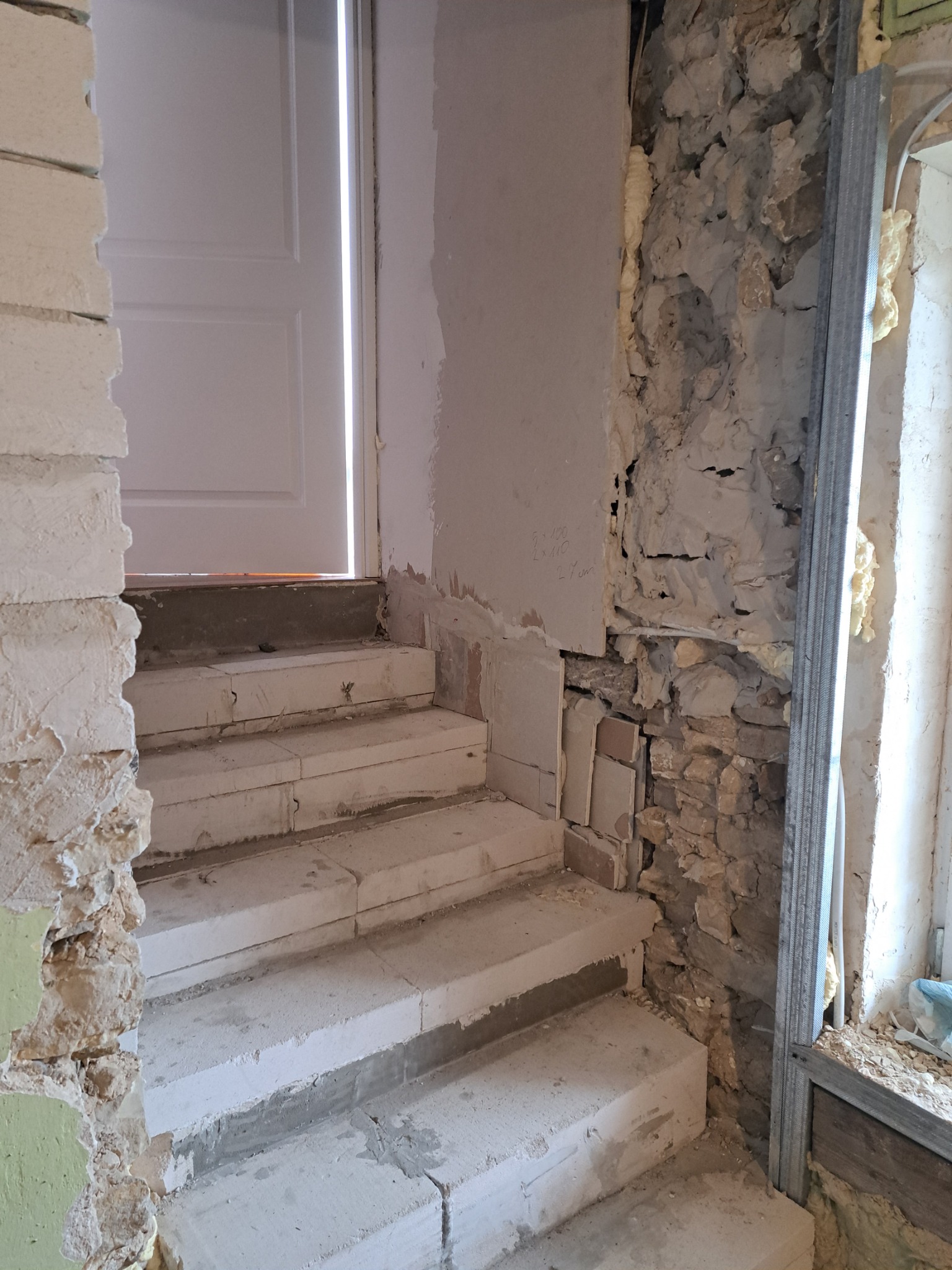 Murowane schody z białych bloczków w trakcie budowy, widoczne surowe ściany z instalacjami i pianką montażową, metalowy profil przy oknie.