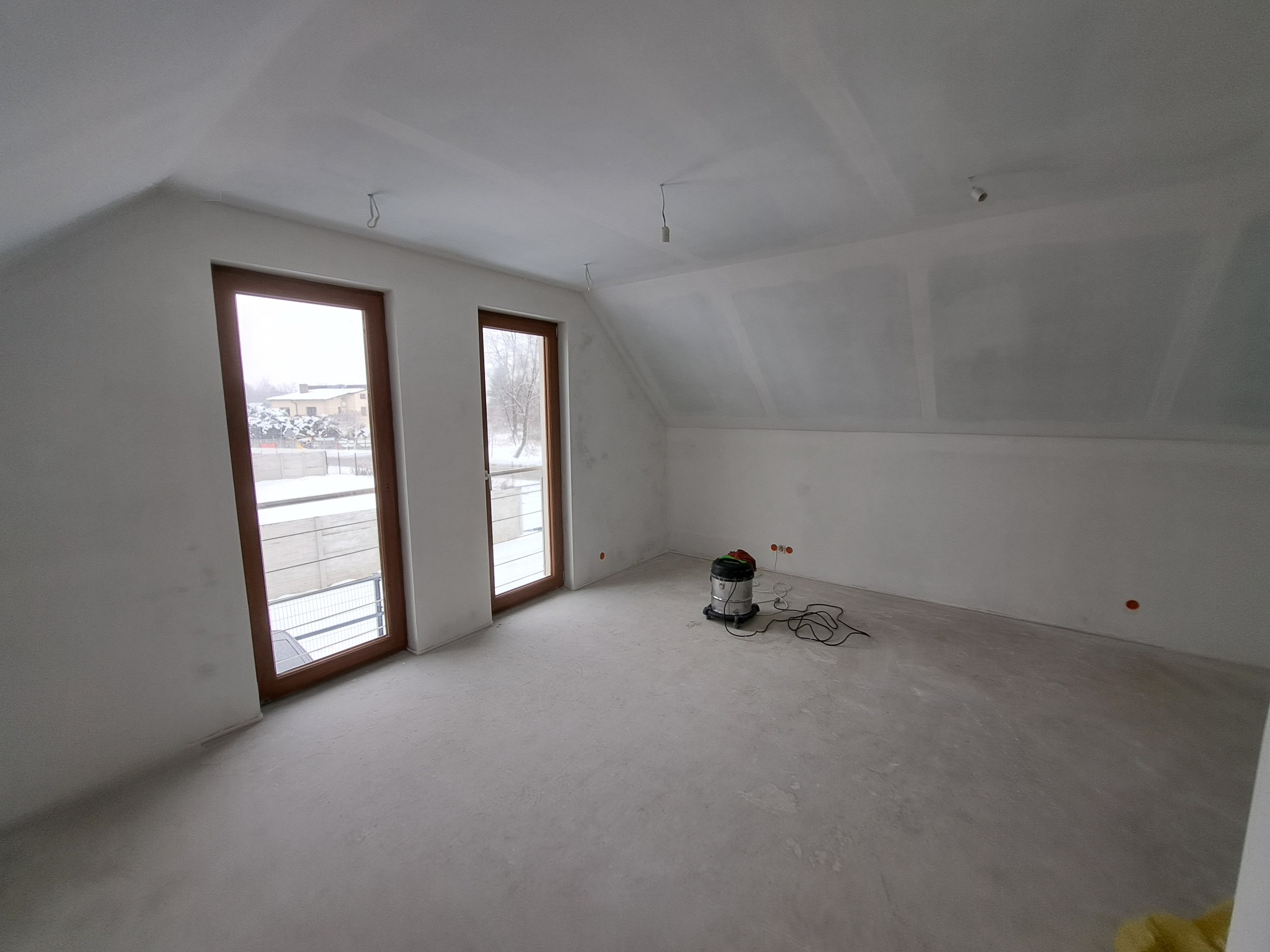 Poddasze po pracach wykończeniowych: świeżo pomalowane ściany, dwa drewniane okna, surowa podłoga i przemysłowy odkurzacz na środku pomieszczenia.