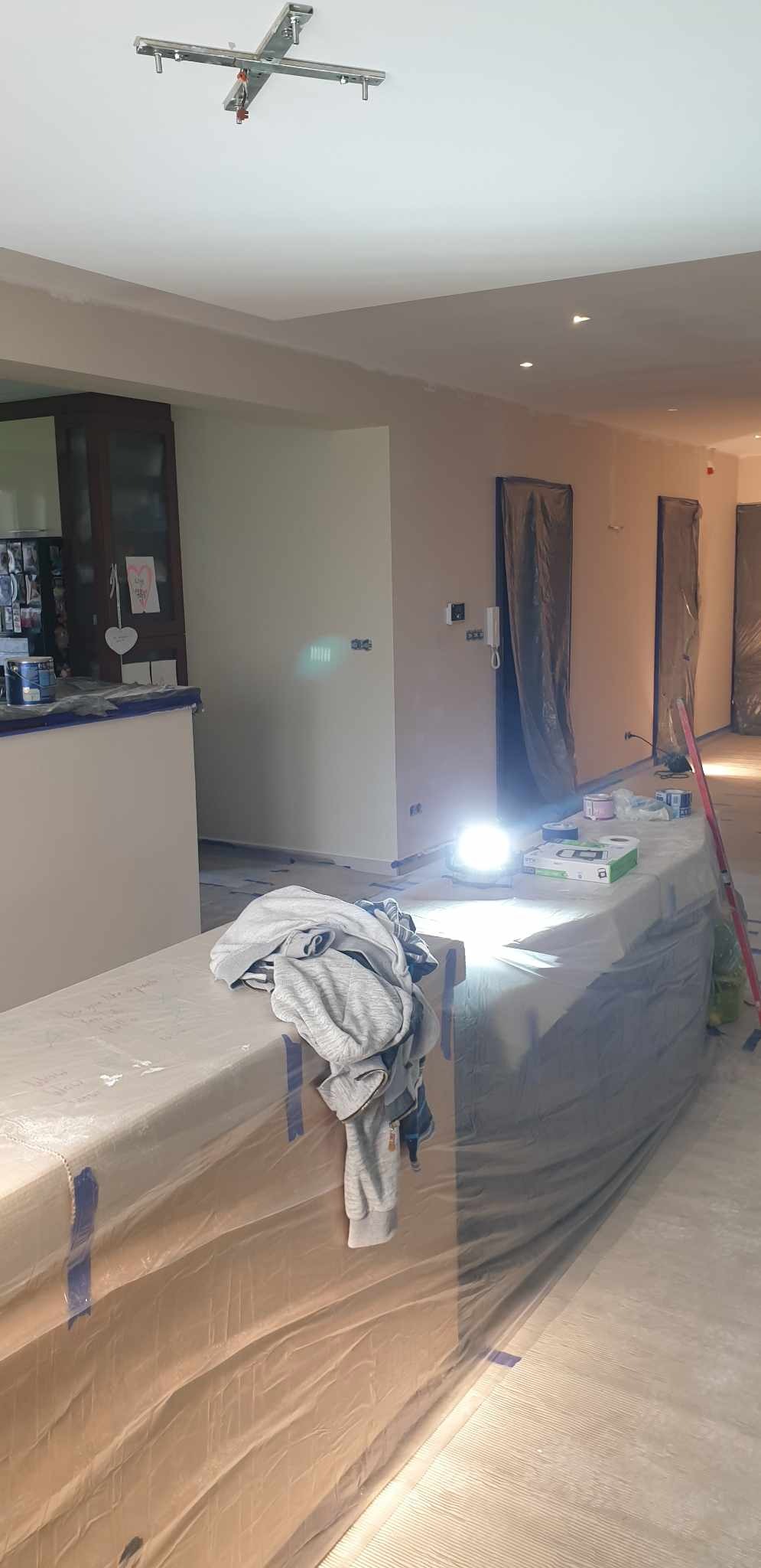 Wnętrze w trakcie malowania: ściany w jasnym odcieniu, zabezpieczone folią meble, rozstawione farby i akcesoria malarskie, widoczne punkty świetlne na suficie i lampa halogenowa oświetlająca...