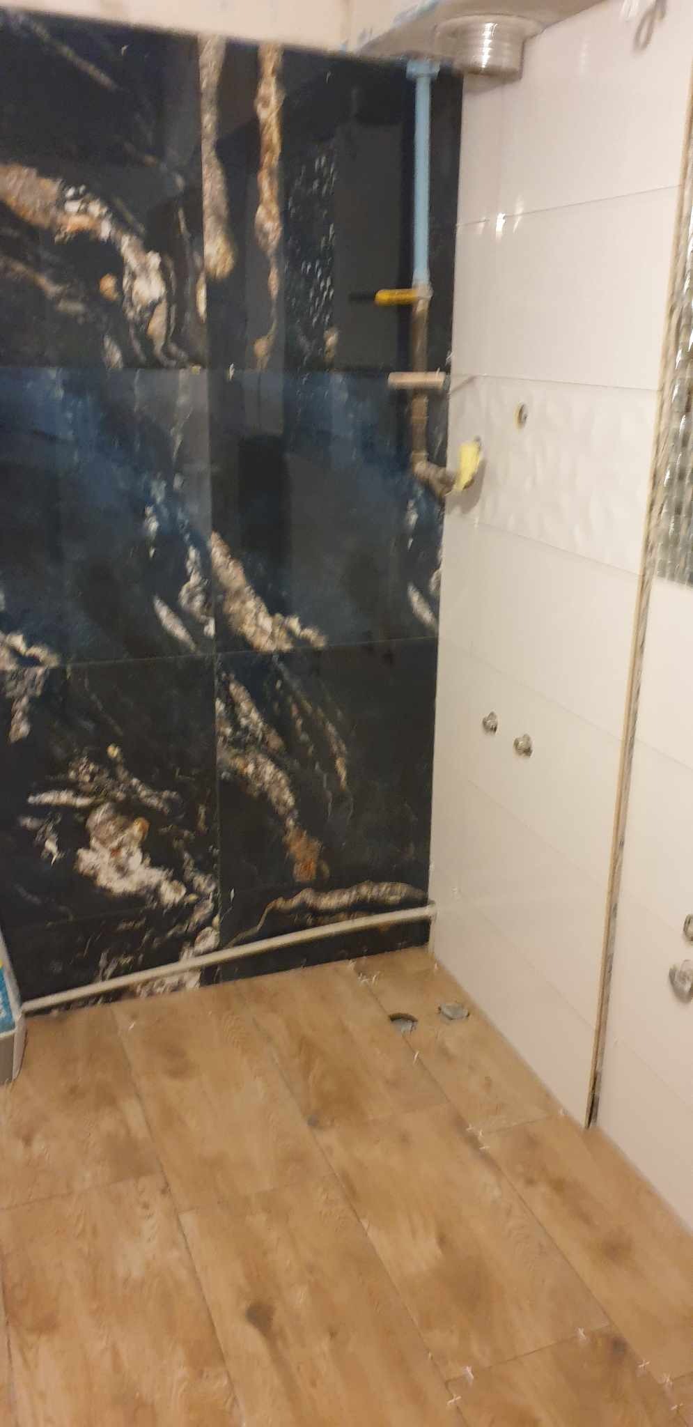Łazienka w trakcie prac wykończeniowych: położone płytki imitujące drewno na podłodze, czarne płytki z wzorem na ścianie, białe płytki na drugiej ścianie, widoczne otwory na rury i instalacje.