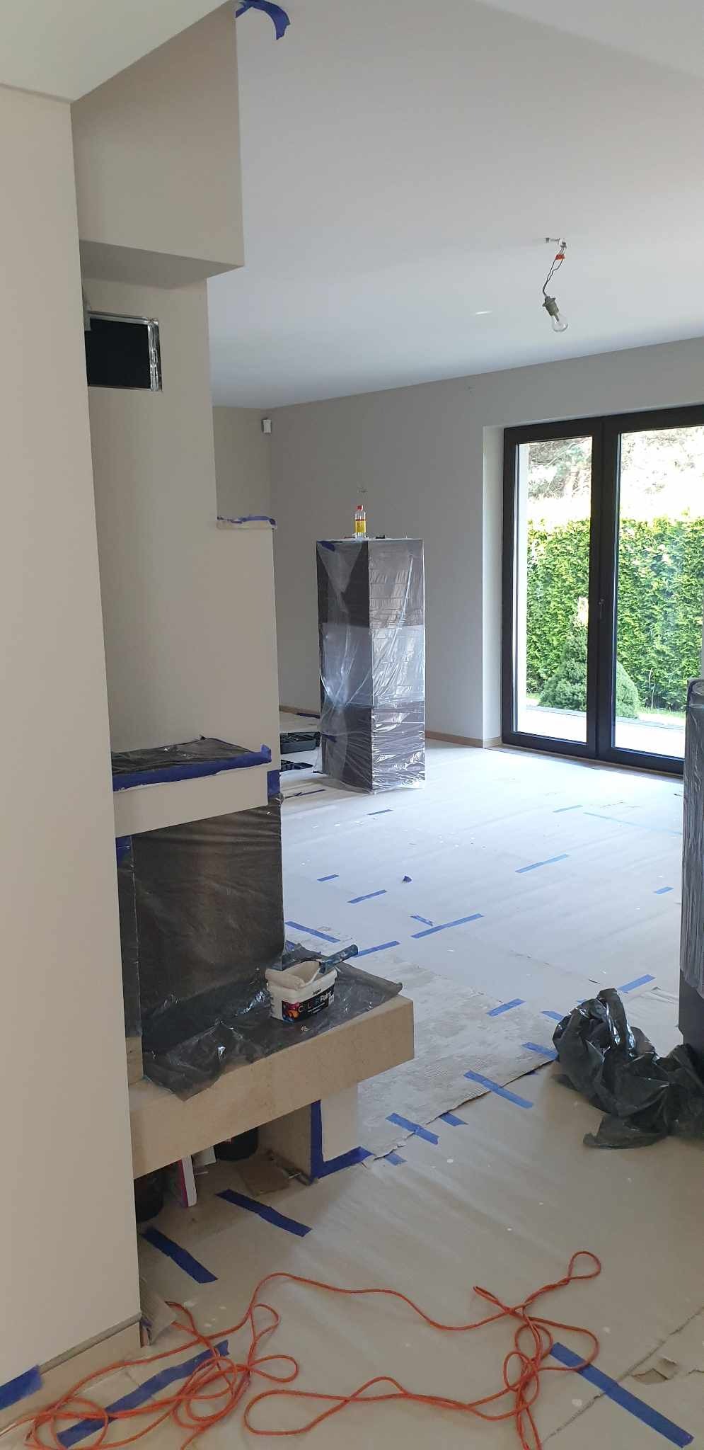 Pokój w trakcie odświeżania: ściany pomalowane na szaro, podłoga zabezpieczona folią i taśmą malarską, meble okryte folią ochronną, widoczny otwarty pojemnik z białą farbą i pędzel na kominku.
