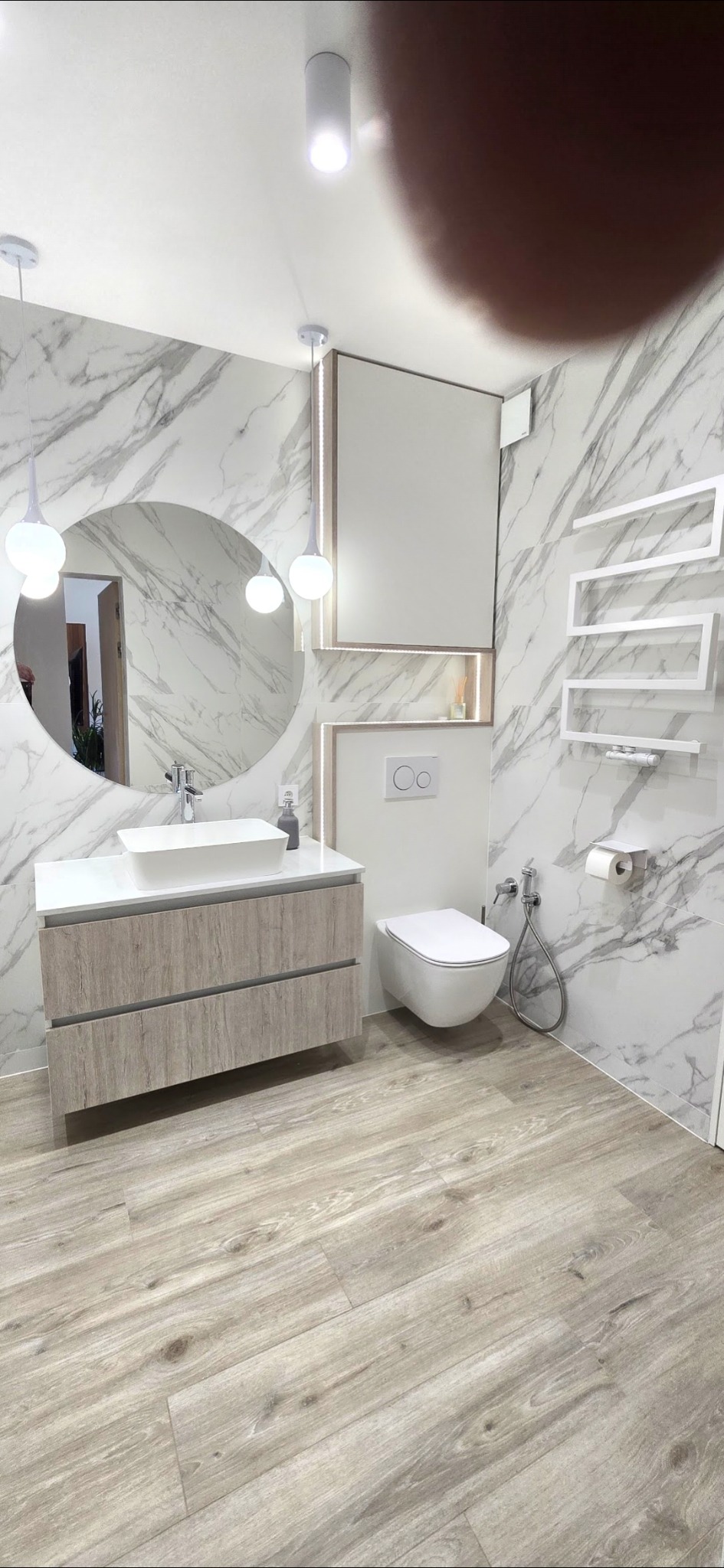 Nowoczesna łazienka z marmurowymi płytkami, okrągłym lustrem z oświetleniem, wiszącą toaletą i grzejnikiem drabinkowym. Jasne drewno na podłodze i szafce pod umywalką.