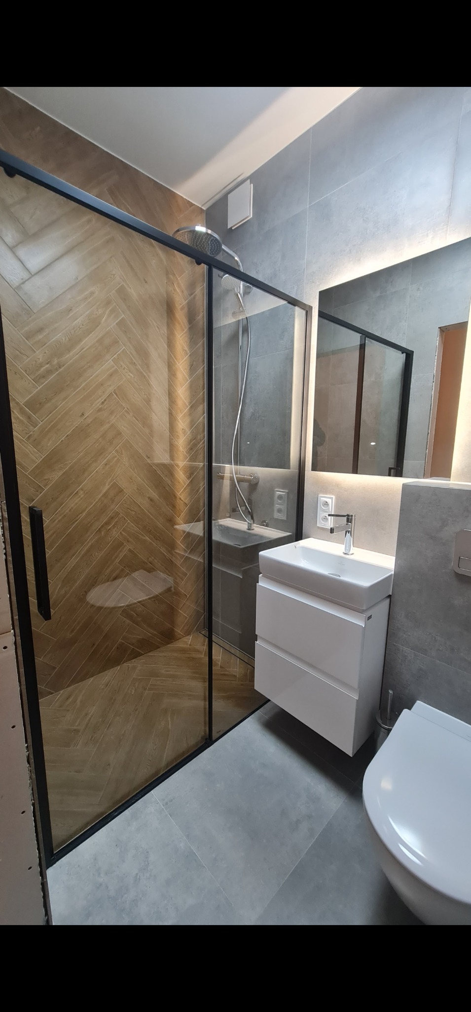 Nowoczesna łazienka z kabiną prysznicową typu walk-in z czarnymi profilami, płytkami ułożonymi w jodełkę wewnątrz prysznica, białą szafką z umywalką i podświetlanym lustrem.