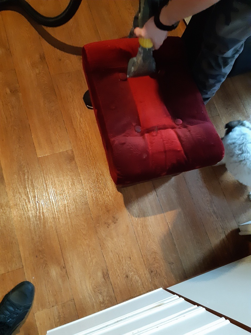 Czerwony, tapicerowany puf czyszczony parowo za pomocą dyszy odkurzacza piorącego, widoczny fragment drewnianej podłogi i czarny wąż odkurzacza.
