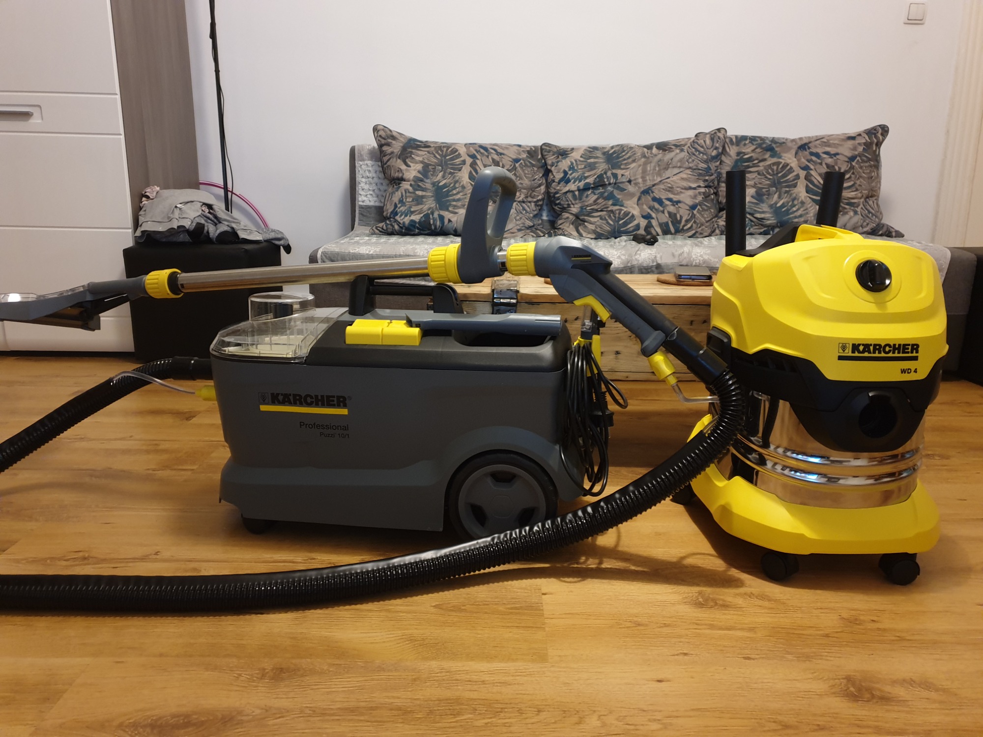 Dwa odkurzacze Kärcher: szary Puzzi 10/1 i żółty WD 4, stojące na drewnianej podłodze w pokoju z sofą w tle.