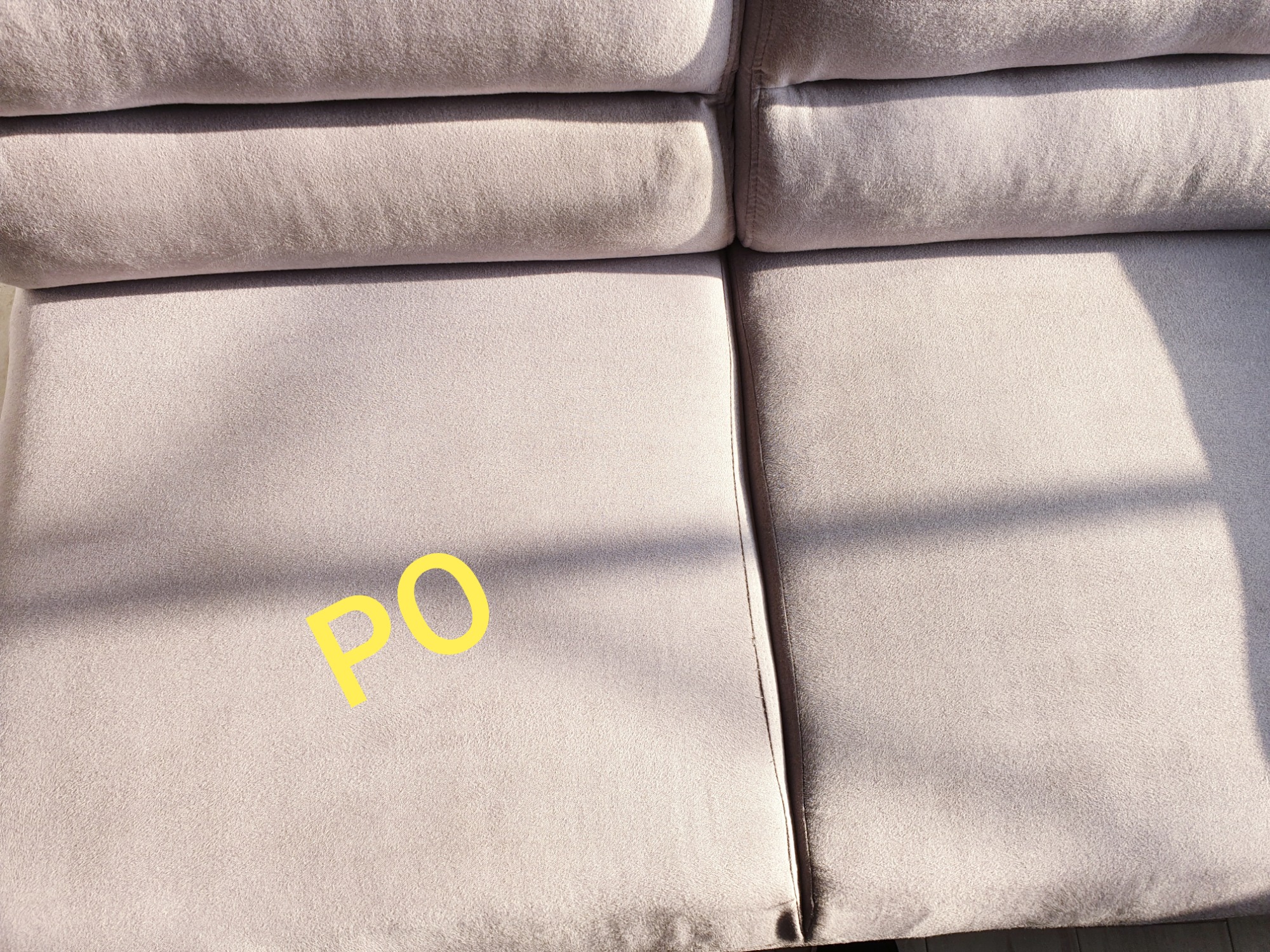 Jasnoszara kanapa z widocznym szwem i napisem 'PO' na siedzisku, oświetlona naturalnym światłem.