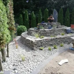 Kaskadowy ogród skalny w trakcie budowy z ułożonymi warstwami kamienia, obsadzony roślinami, z widocznym pracownikiem w kucki na szczycie i taczką w tle, otoczony tujami i wysypany białym żwirem.