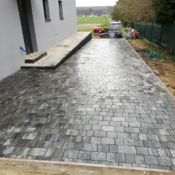 Nowo ułożona, szara kostka brukowa na podjeździe domu jednorodzinnego. Widoczne palety z kostką w tle, zielony trawnik i drzewa.