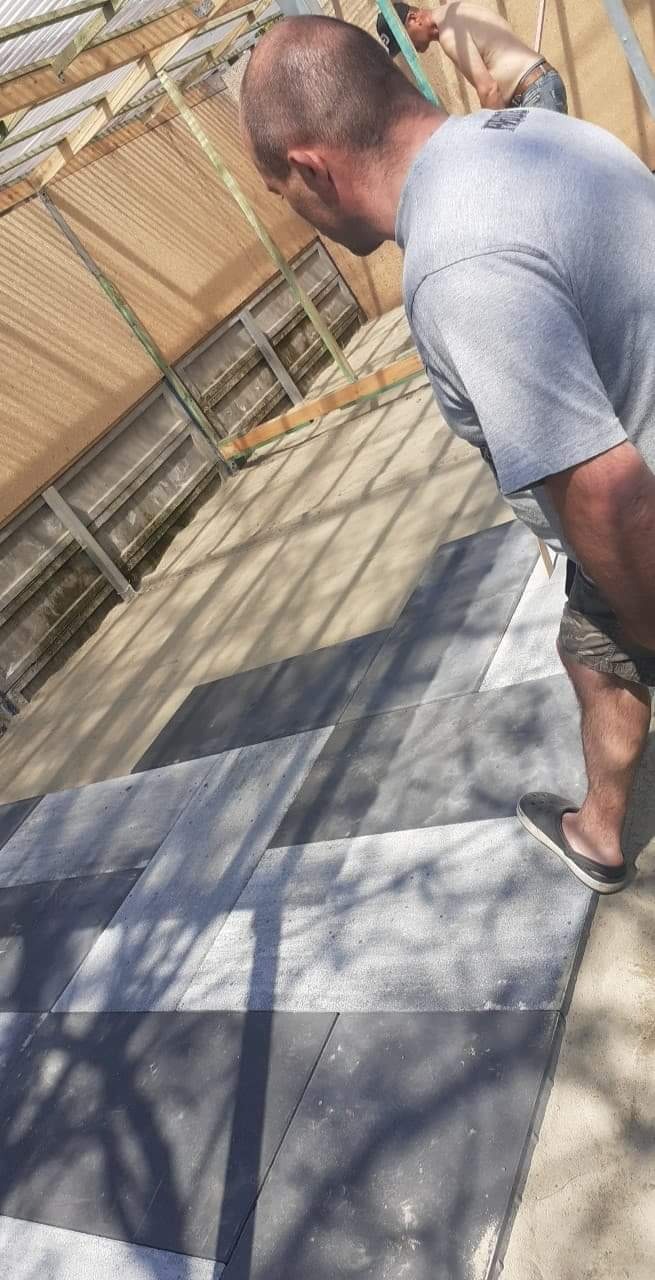 Dwóch mężczyzn układa szaro-białą kostkę brukową w zygzak na piasku pod zadaszeniem z widocznymi elementami konstrukcyjnymi.