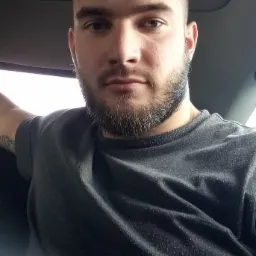 Mężczyzna z brodą w szarej koszulce siedzący w samochodzie, widoczny tatuaż na ramieniu.