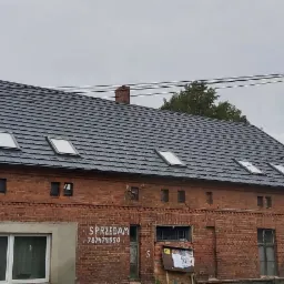 Ciemnoszary dach z dachówki ceramicznej na starym, ceglanym budynku z czterema oknami dachowymi i kominem. Na ścianie napis 'Sprzedam' z numerem telefonu.