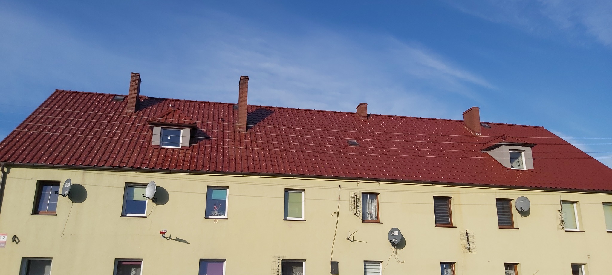 Czerwony dach wielorodzinnego budynku z kilkoma kominami i oknami dachowymi, widok z dołu na tle błękitnego nieba.