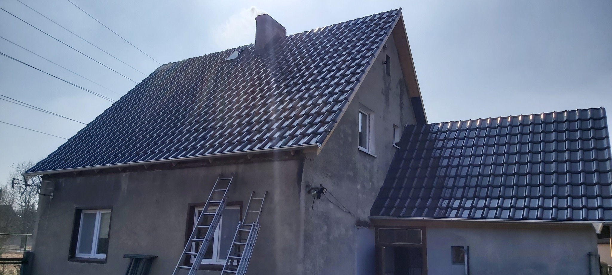 Dom z nowym, błyszczącym, ciemnym dachem ceramicznym, widoczne dwie drabiny opierające się o ścianę, komin z lekkim dymem.